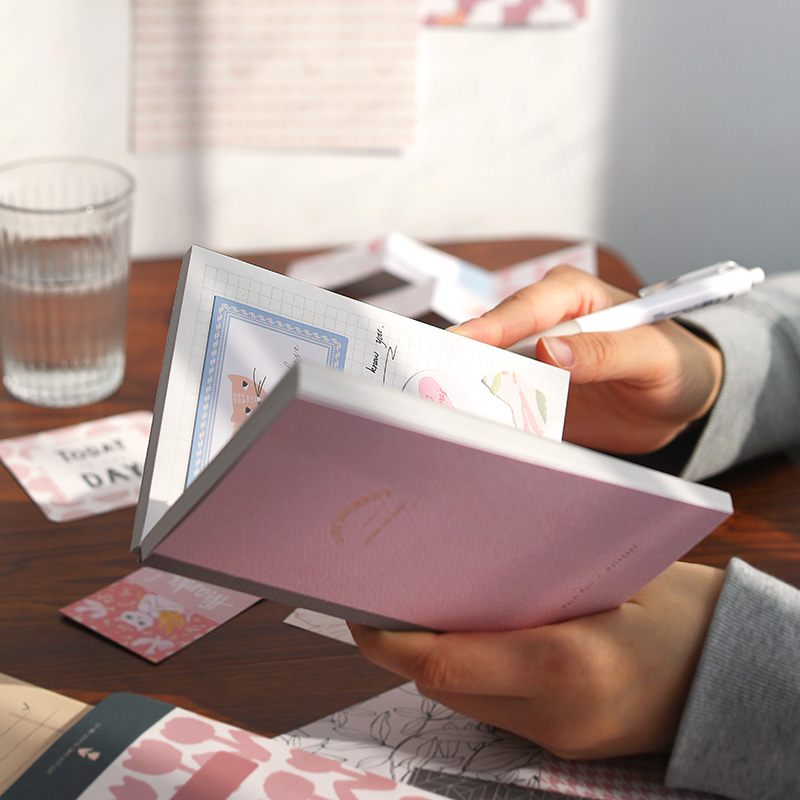 Person holding a pink notebook with stationery items on a desk about to write inside