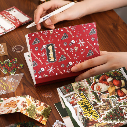 Person holding a red Christmas-themed gift box with decorative stickers on a wooden surface.