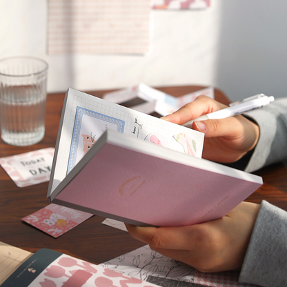 Person holding a pink notebook with stationery items on a desk about to write inside